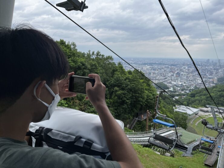 リフトの上から札幌の街並みをスマートフォンで撮影する生徒の後ろ姿