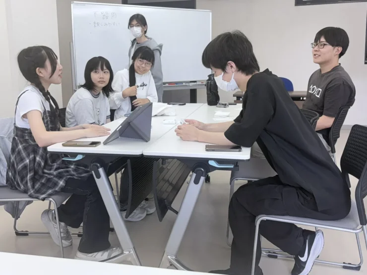 ホワイトボードの前でテーブルを囲み、学年をこえて仲間の話に耳を傾ける生徒たち